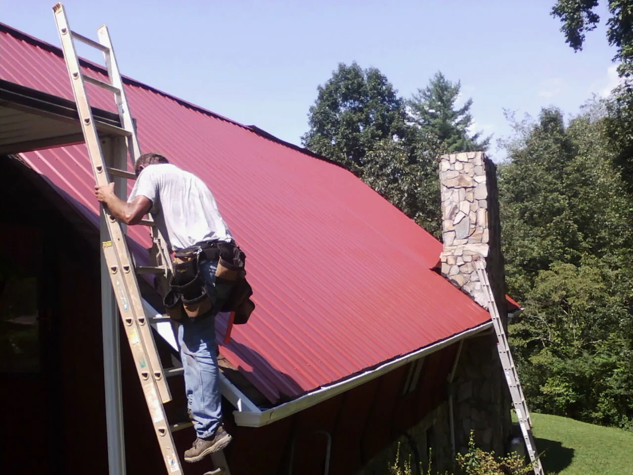 Roofer on a ladder beside a red metal roof and stone chimney — Soffit & Fascia Repair by Roofing Brothers in Port William